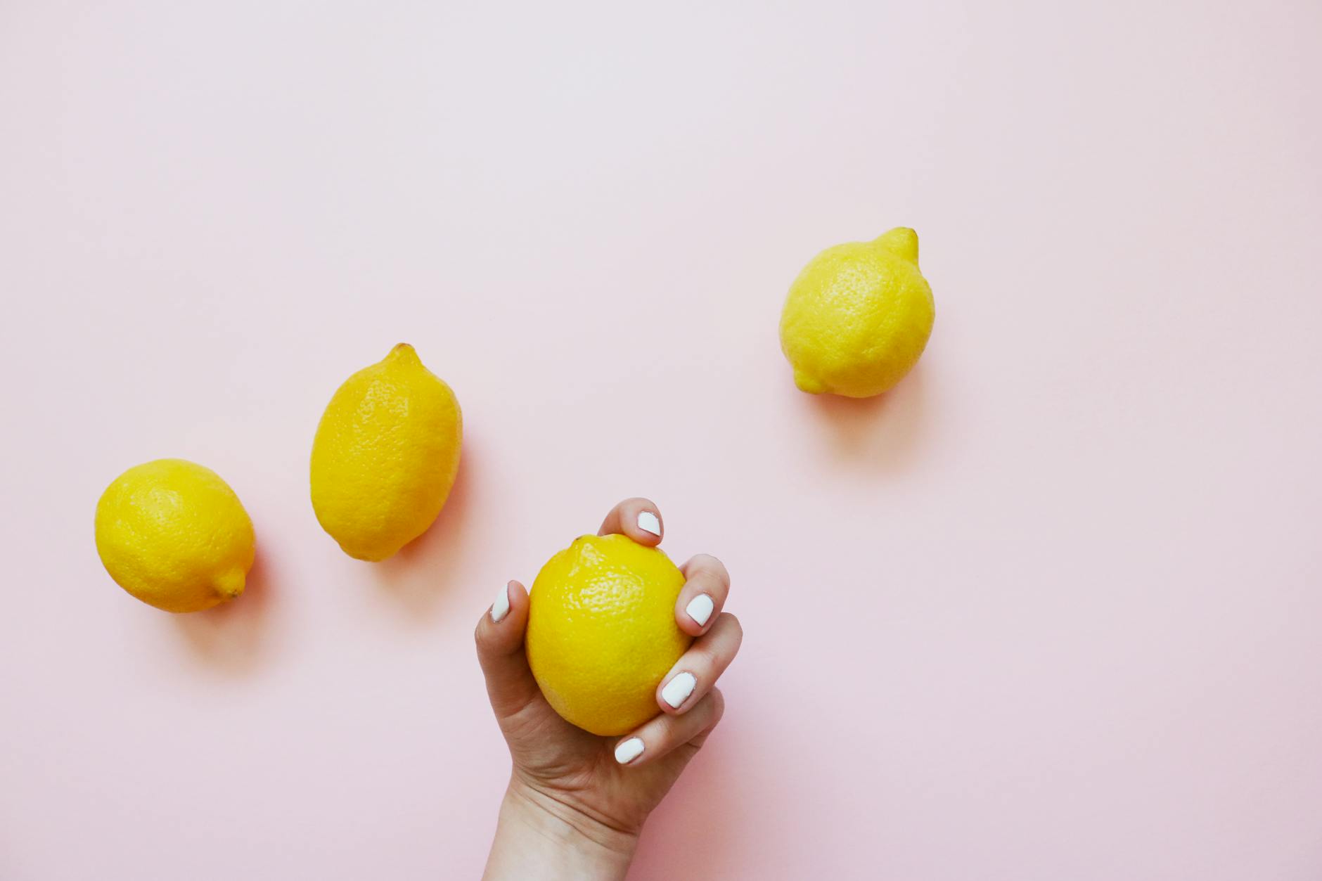 Fotografía conceptual de una mano sosteniendo un limón sobre fondo rosa suave, rodeada de otros limones.