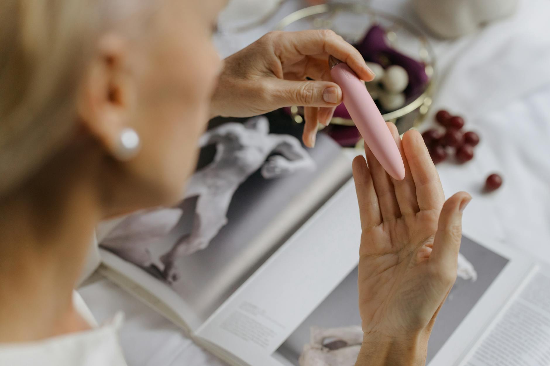 Primer plano de una mujer sosteniendo un vibrador rosa, con libros de arte de fondo, enfatizando temas íntimos y personales.